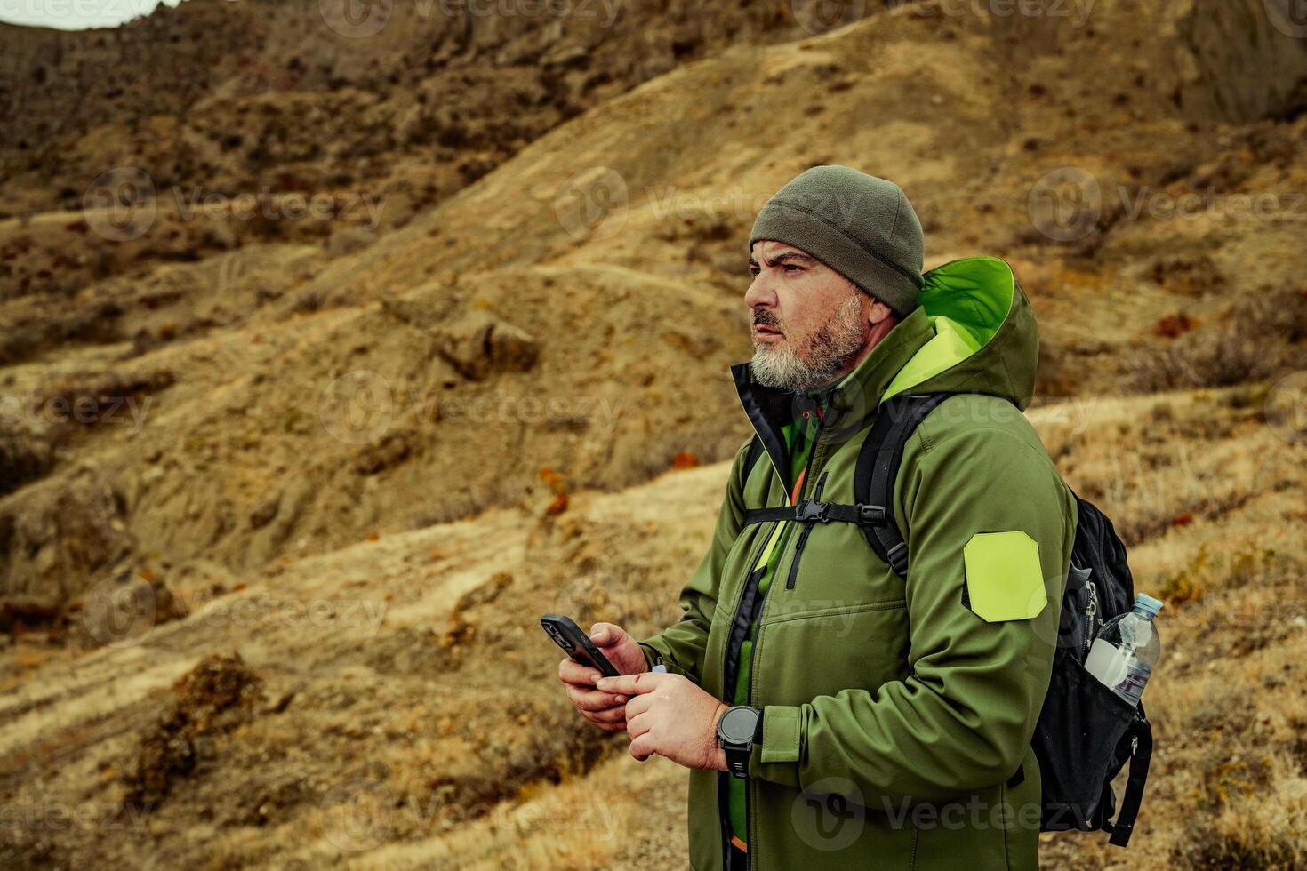Man standing on rocky terrain in rugged outdoor gear while checking his phone during an adventure in nature photo