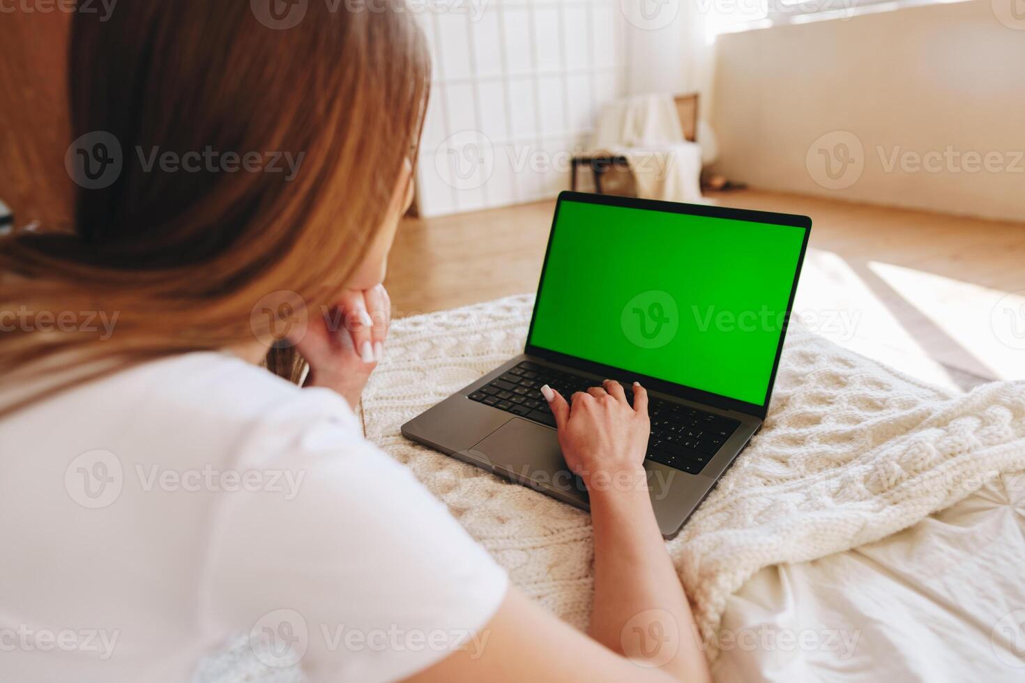 Young person using a laptop with a green screen while sitting on a bed in a cozy room during the day photo