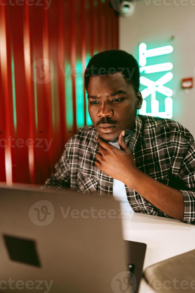 Focused young man working on laptop in modern workspace with neon exit sign during evening hours photo