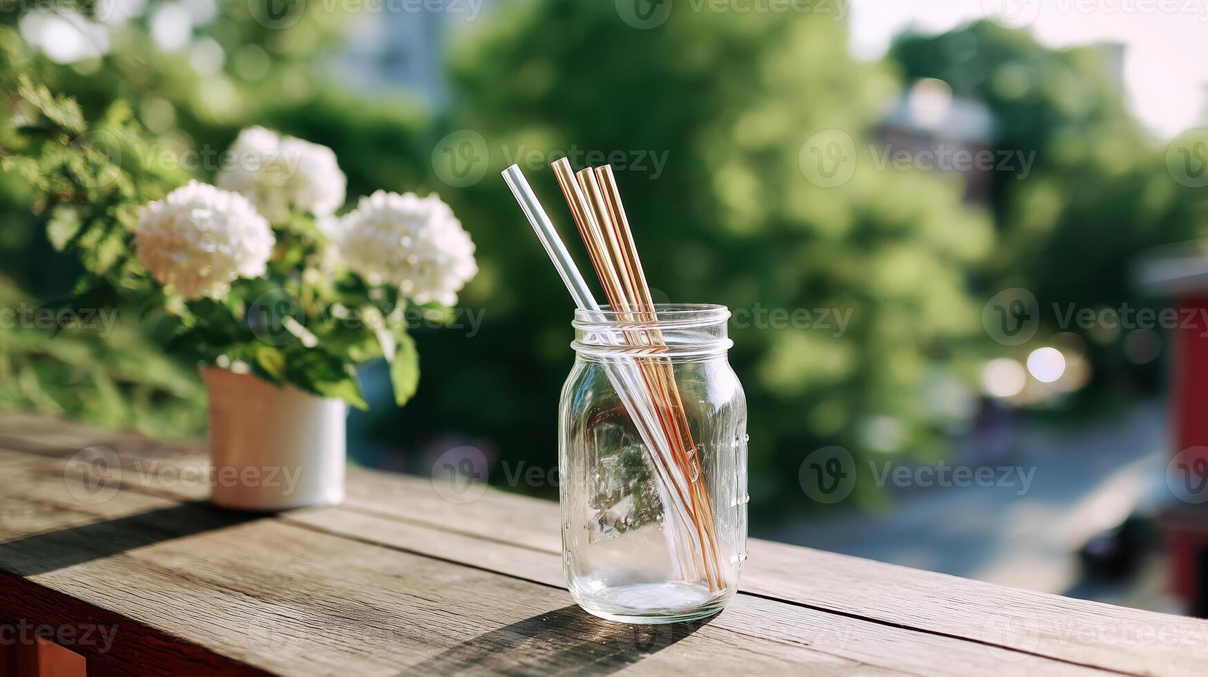 Glass jar holds reusable straws on wooden railing with potted flowers in a sunny outdoor setting photo