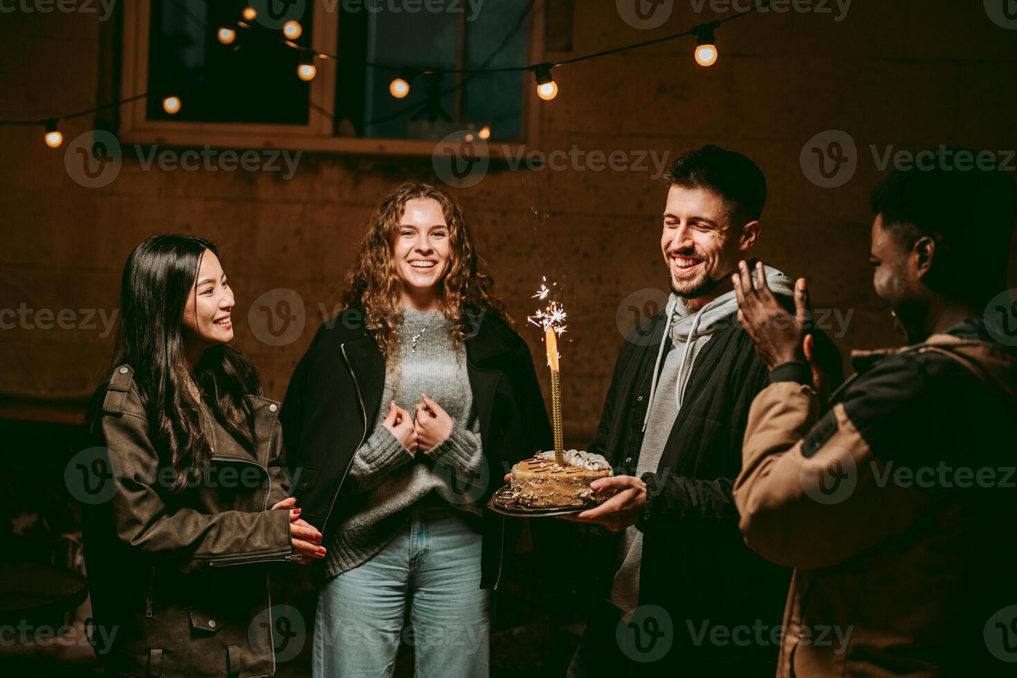 Group of friends celebrating a birthday outdoors at night with lights and a cake photo