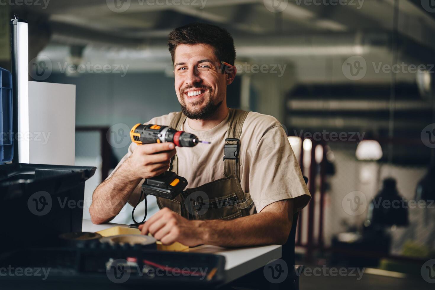 Smiling craftsman holds drill while working in workshop during the day photo