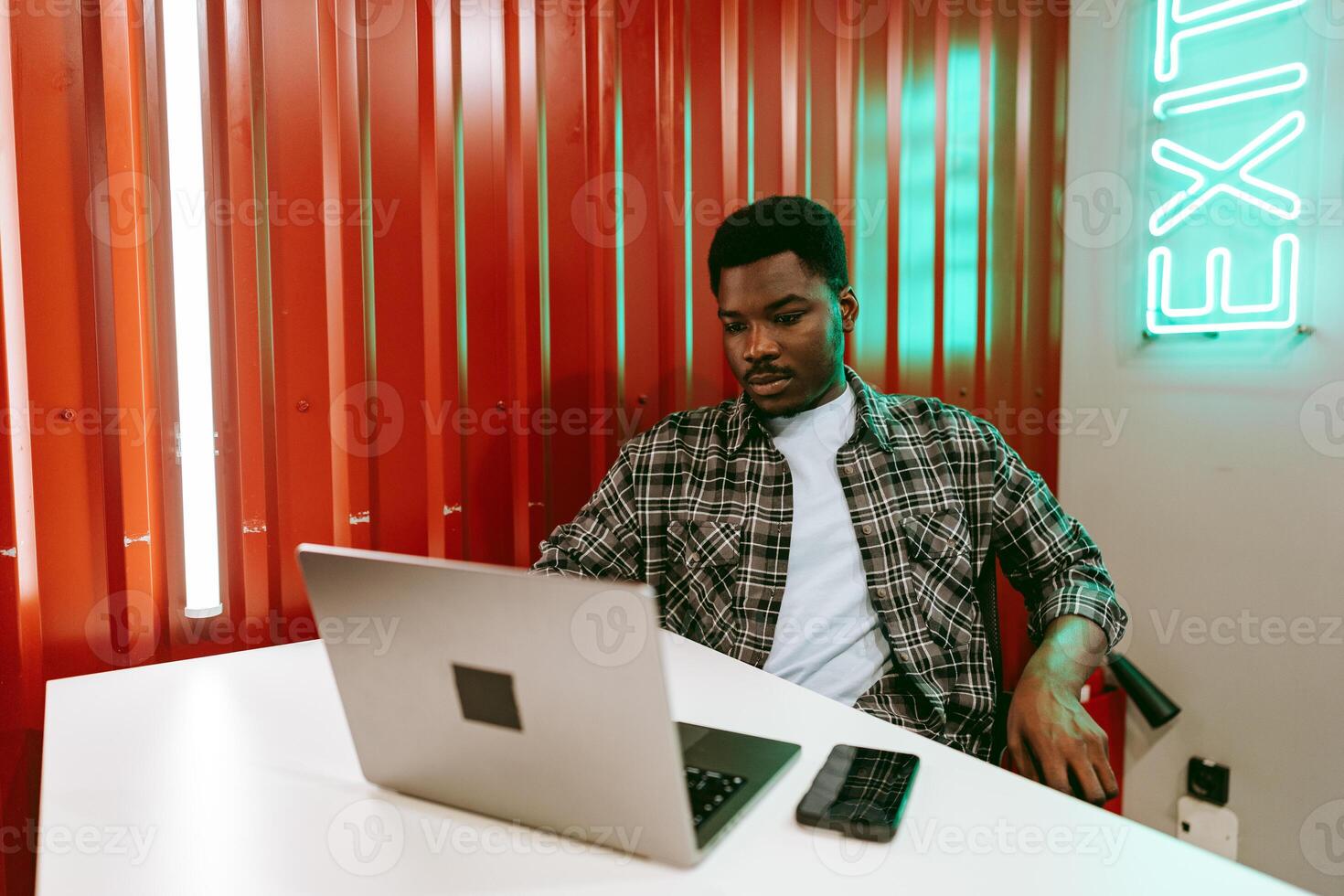 Young man working on laptop in modern coworking space with vibrant red walls and green neon sign photo