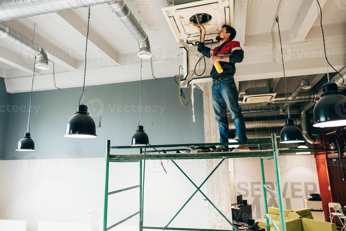 Worker performs maintenance on HVAC system while standing on scaffolding in an office space during the daytime photo