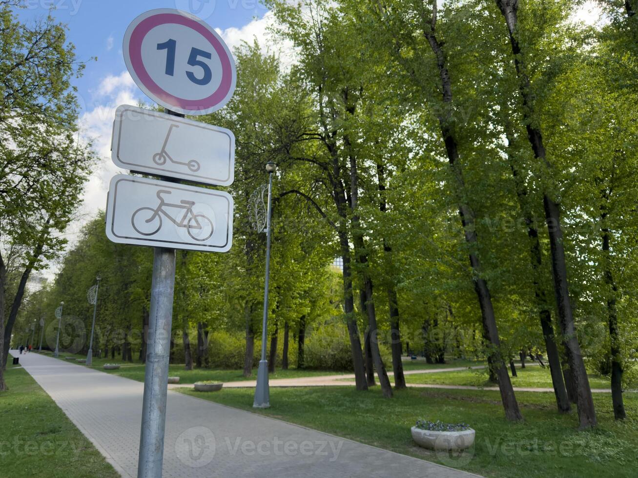 Speed limit sign for bicycles and scooters on a park pathway surrounded by tall green trees. Urban park infrastructure promoting safety and eco-friendly transportation photo