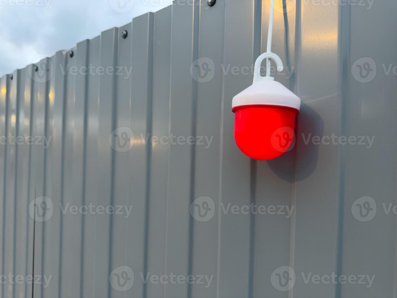 Close up of red warning light mounted on gray metal fence at construction site during twilight. The glowing lamp adds contrast and draws attention to safety and hazard awareness in public spaces photo