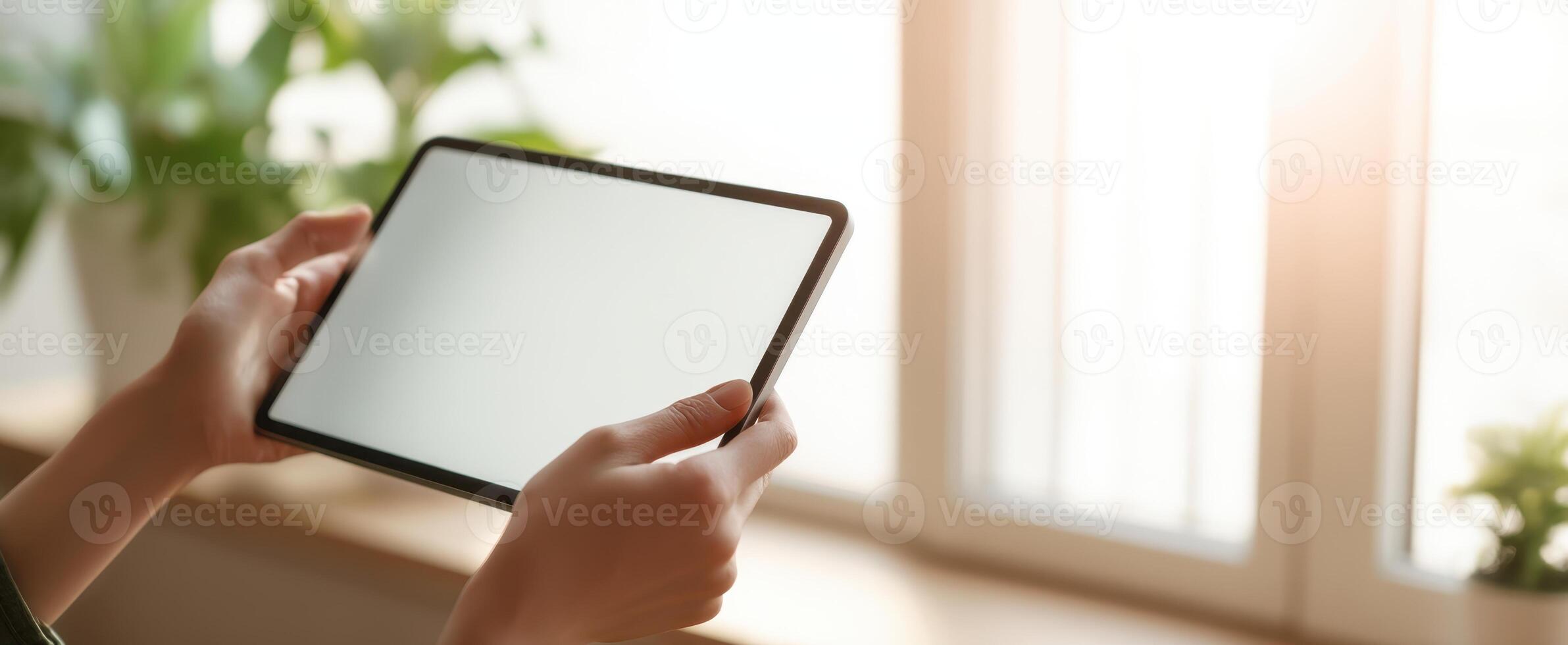 The tablet held in hands against a sunlit window with plants. photo