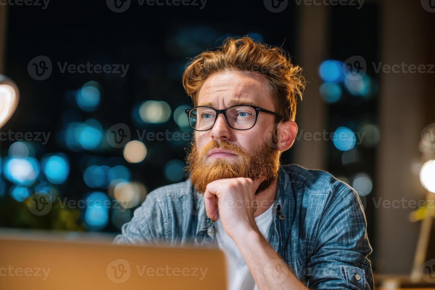 A bearded man stares thoughtfully at a laptop screen at nighttime photo