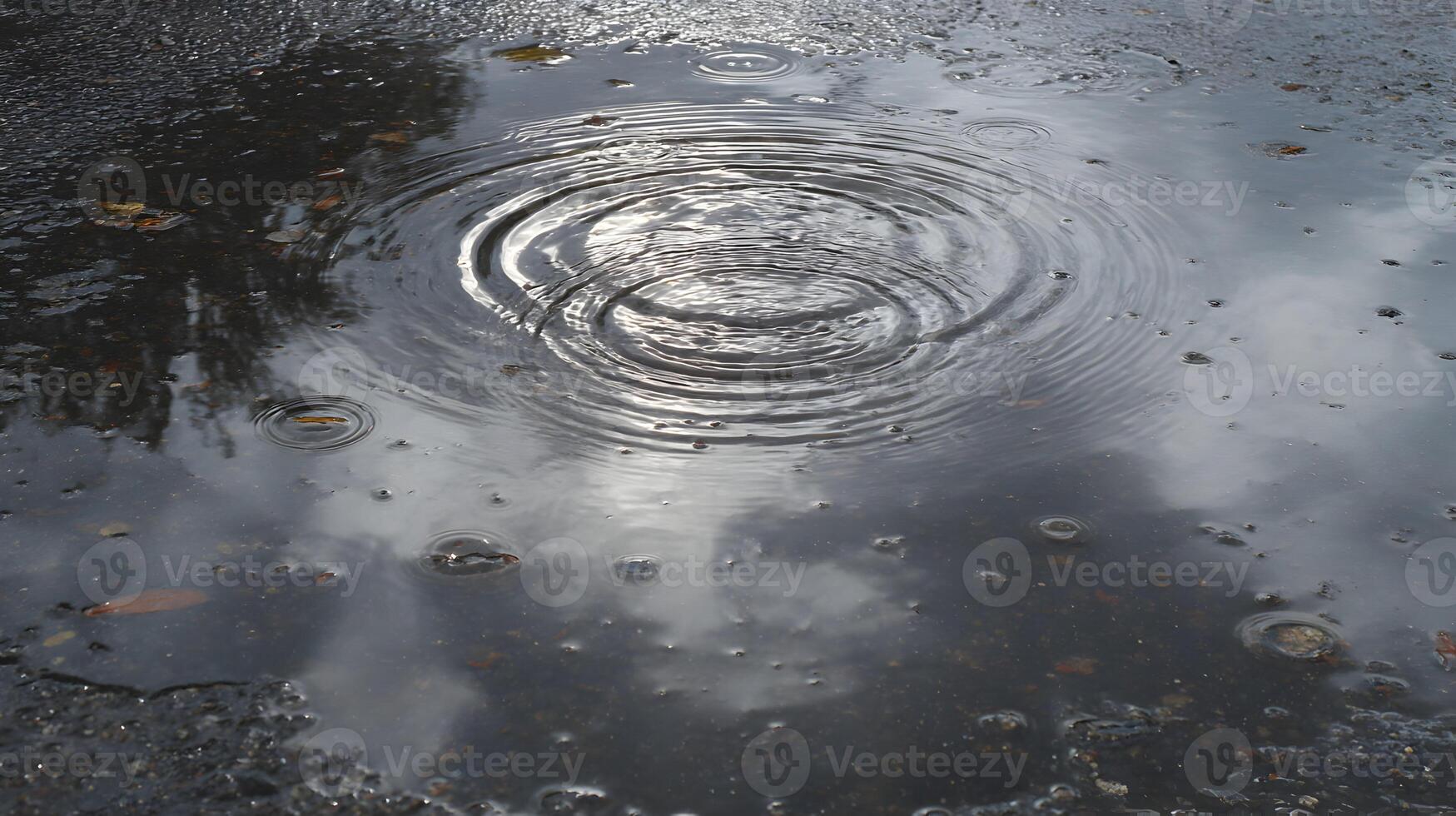 Raindrops falling create ripples in a puddle reflecting the sky adding texture to the asphalt surface photo