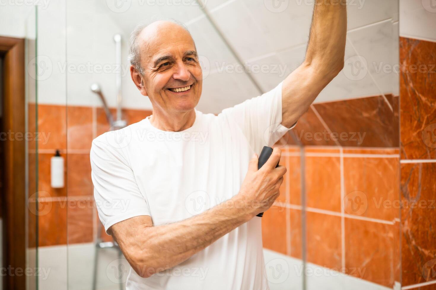 Portrait of elderly man with gray hair applying deodorant in front of mirror while standing in bathroom. He is enjoying his fresh morning ritual with scented body spray. photo