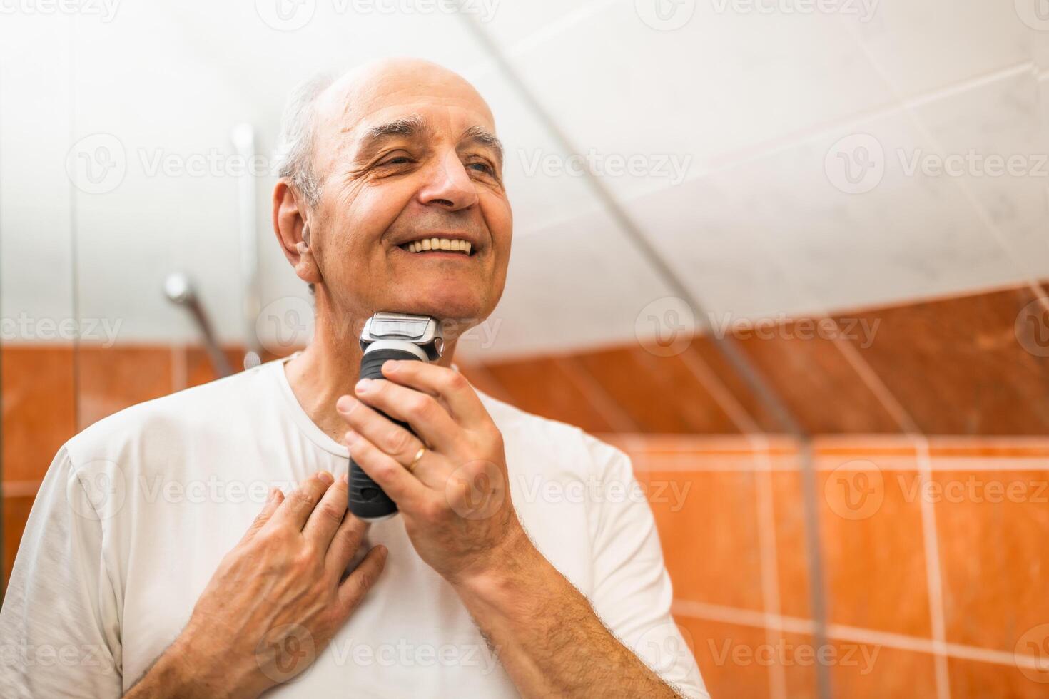 Handsome senior man smiling while shaving his beard with electric razor in front of bathroom mirror, focusing on hygiene, beauty and personal care. photo