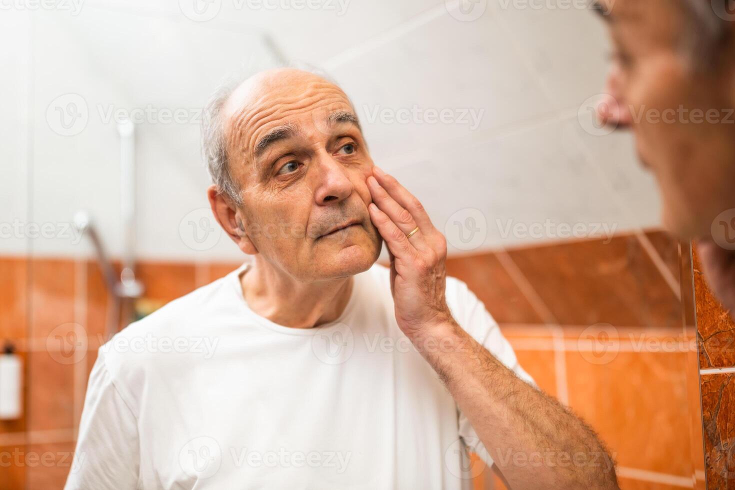 Displeased senior man standing in front of mirror and looking at his eye bags. He is visibly tired and worried while analyzing his reflection in bathroom setting. photo