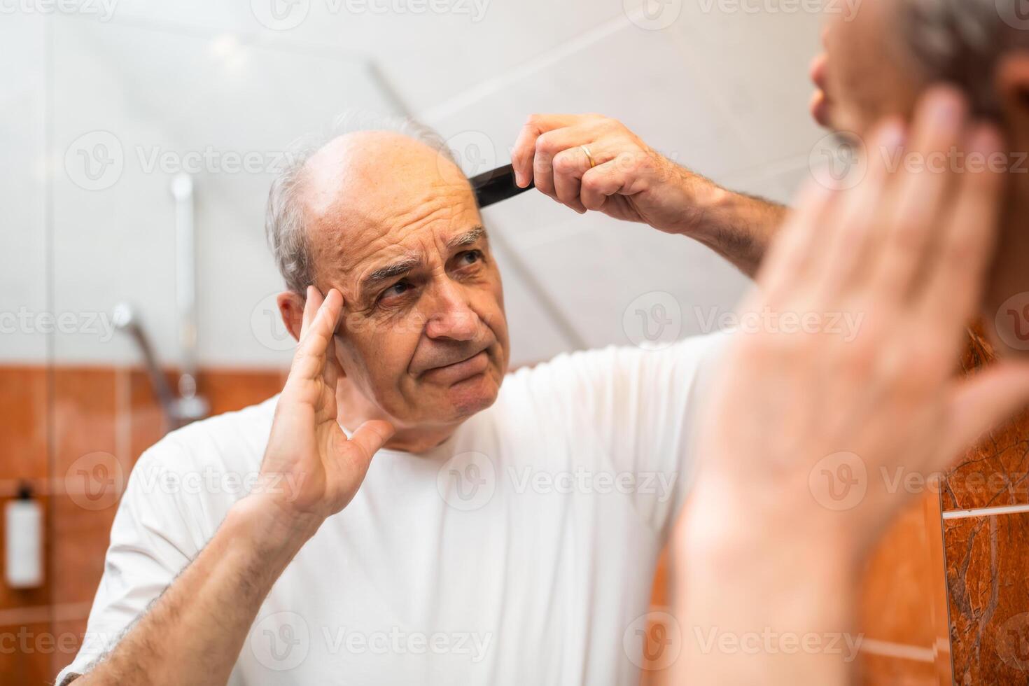 Aged male in his 60s feeling displeased while combing his hair in front of mirror in bathroom. He is expressing emotional impact of aging and hair loss. photo