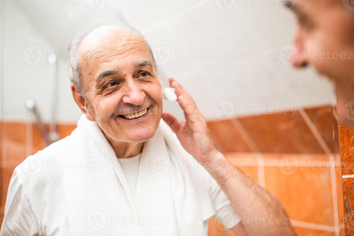Portrait of confident elderly man with towel around his neck applying shaving cream while looking at his reflection in bathroom mirror, promoting masculinity, hygiene and active senior lifestyles. photo