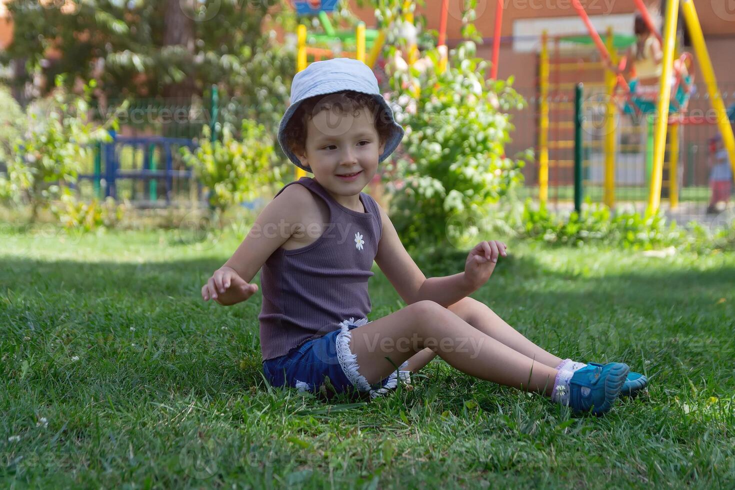 A child in a panama hat sits on the lawn near the playground photo