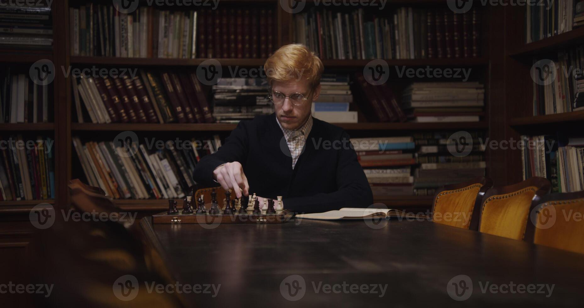 A young man of European appearance, dressed in a business suit, is studying with concentration to play chess alone, using a textbook, sitting at a table photo