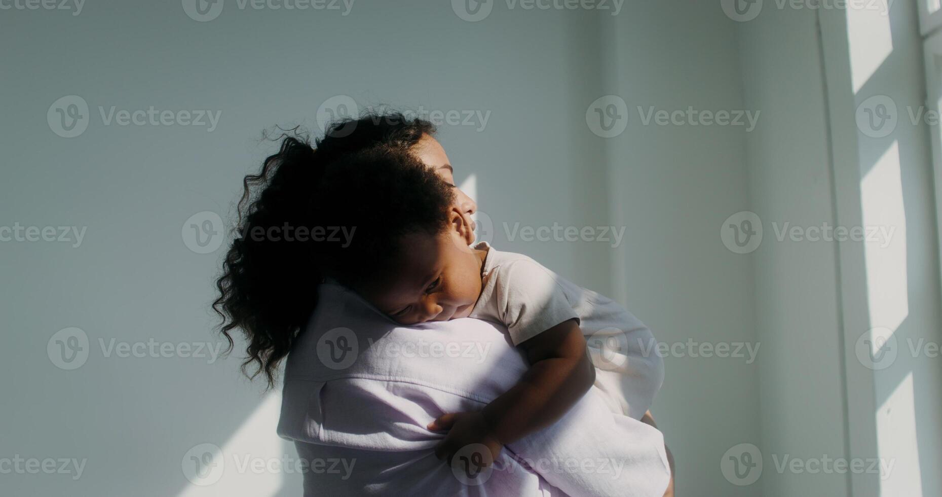 An African-American woman dressed in pastel-colored clothes holds her little daughter in her arms photo