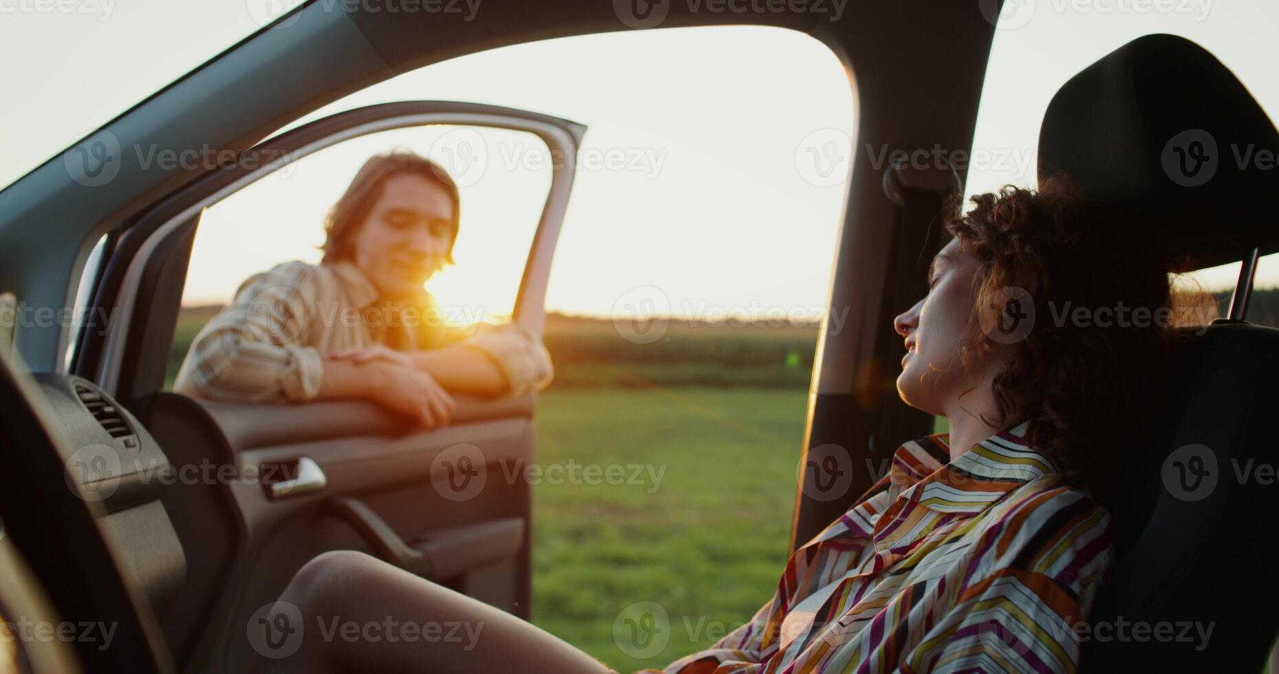A woman is sitting in a car, a man is standing near the open door, they are smiling and talking. photo