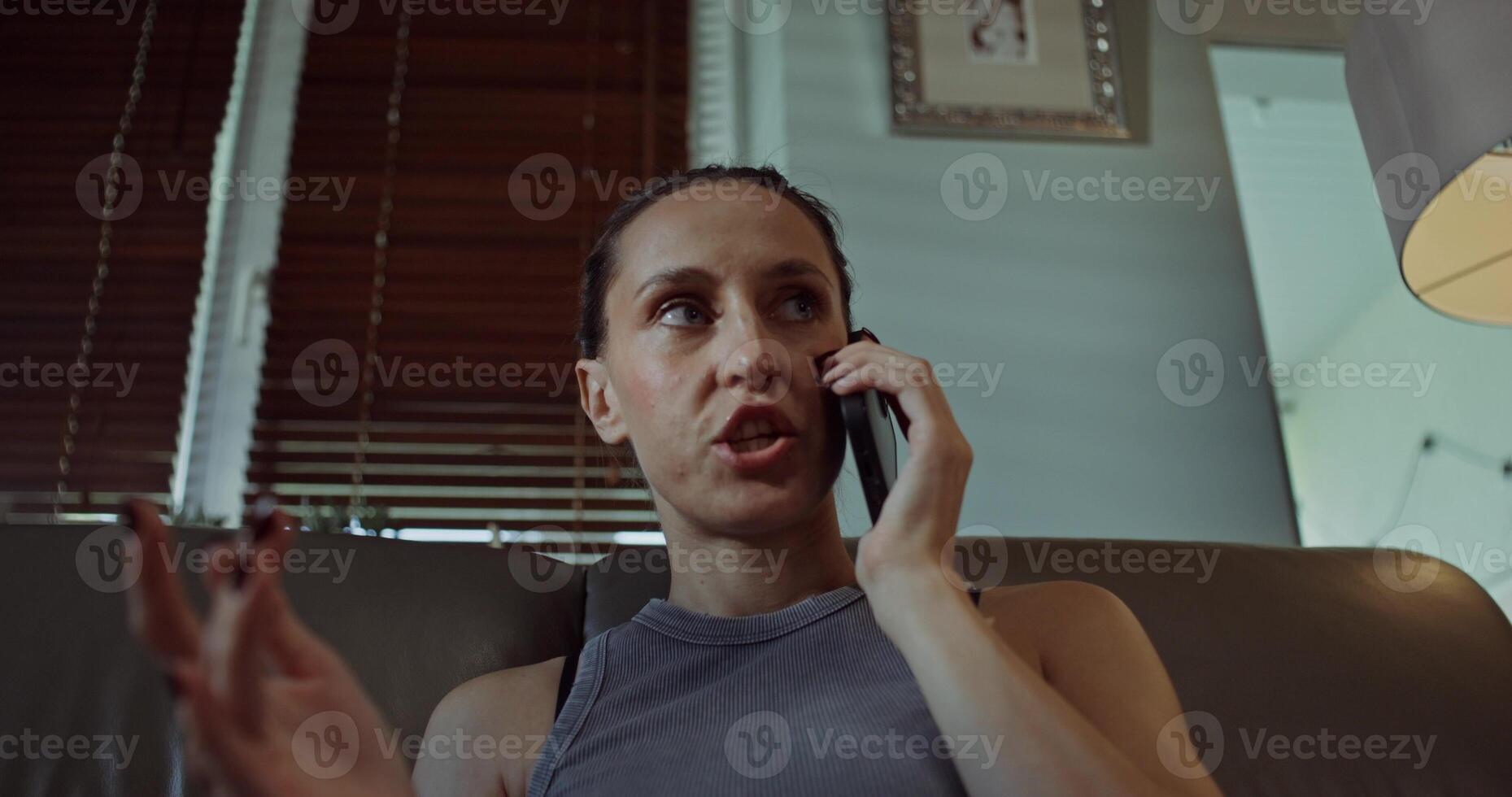 A woman is talking on a mobile phone while sitting on the couch at home. She looks angry. photo