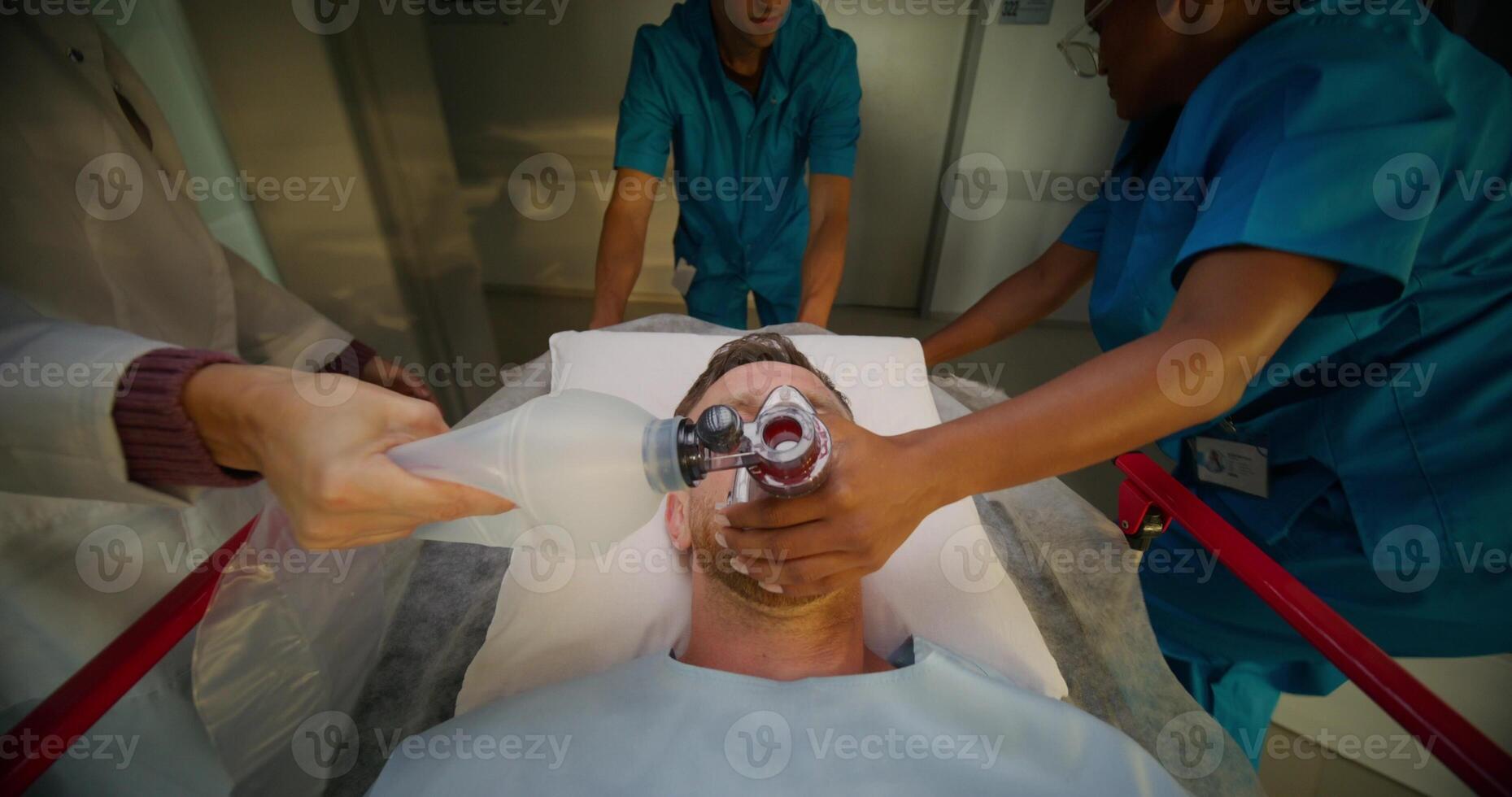 A man is lying on a medical gurney, he is being taken through the hospital corridor. A nurse holds an oxygen mask photo