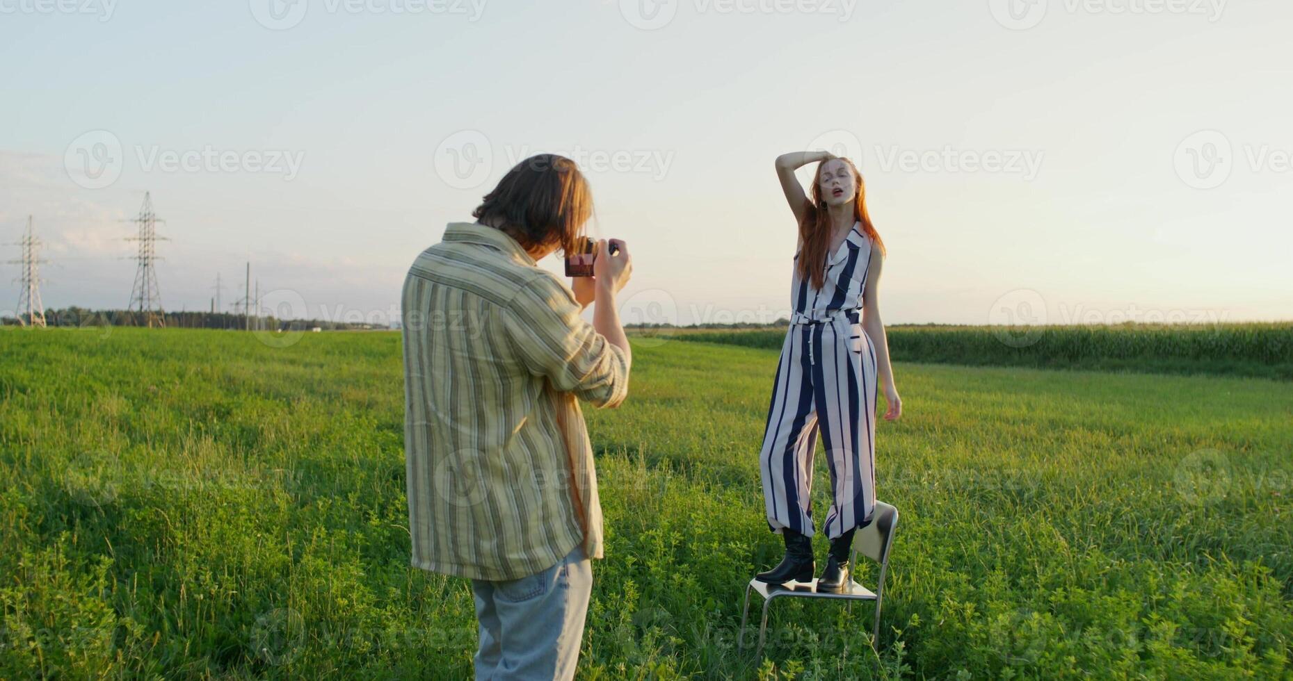A man takes pictures of a young woman at sunset in a field. A woman poses standing on a chair photo