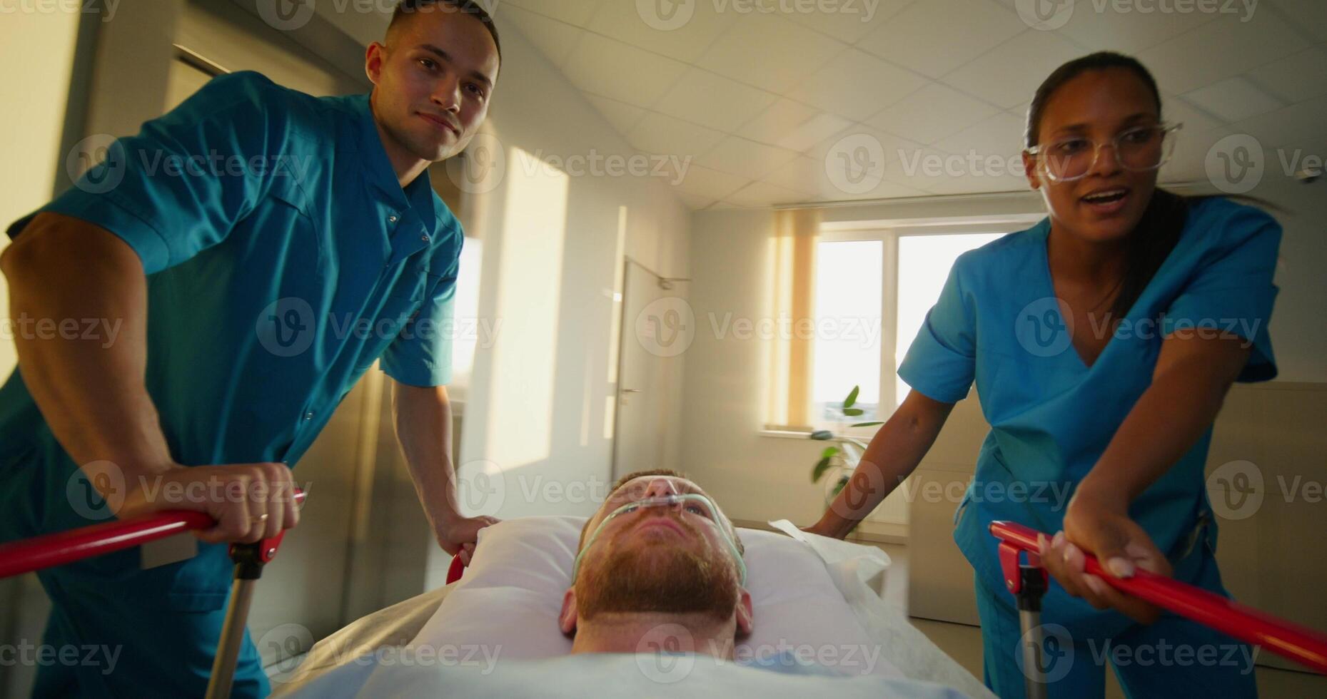 A man with an oxygen tube in his nose is lying on a medical gurney. The orderlies talk and smile as they take the patient through the hospital corridor photo