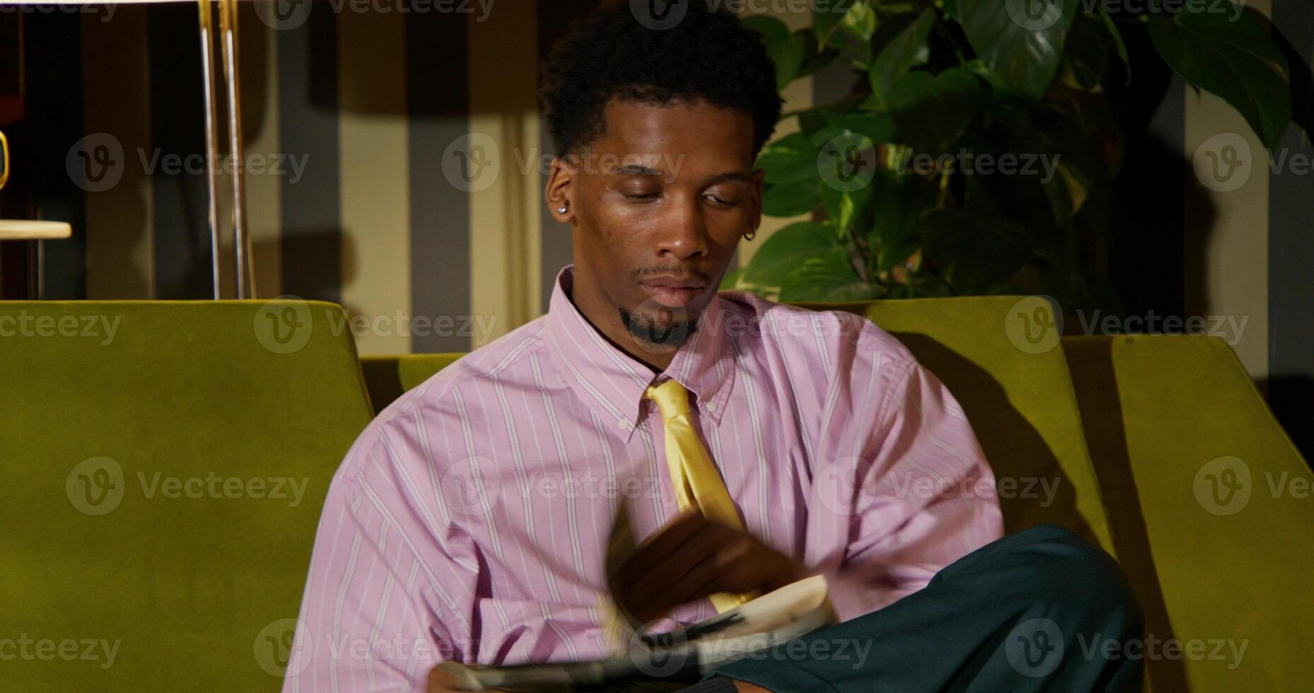 A man is reading a newspaper with a concentrated look while sitting on the couch at home. His wife is on the phone sitting on the floor next to him. photo