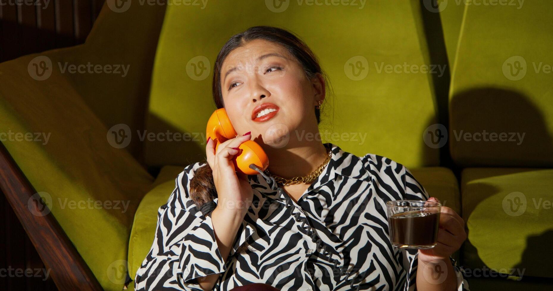 A woman smiles while talking on a wired phone. She is sitting on the floor next to a sofa where a man is reading a newspaper photo