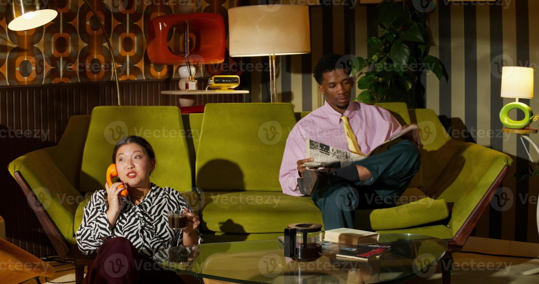 A woman smiles while talking on a wired phone. She is sitting on the floor next to a sofa where a man is reading photo