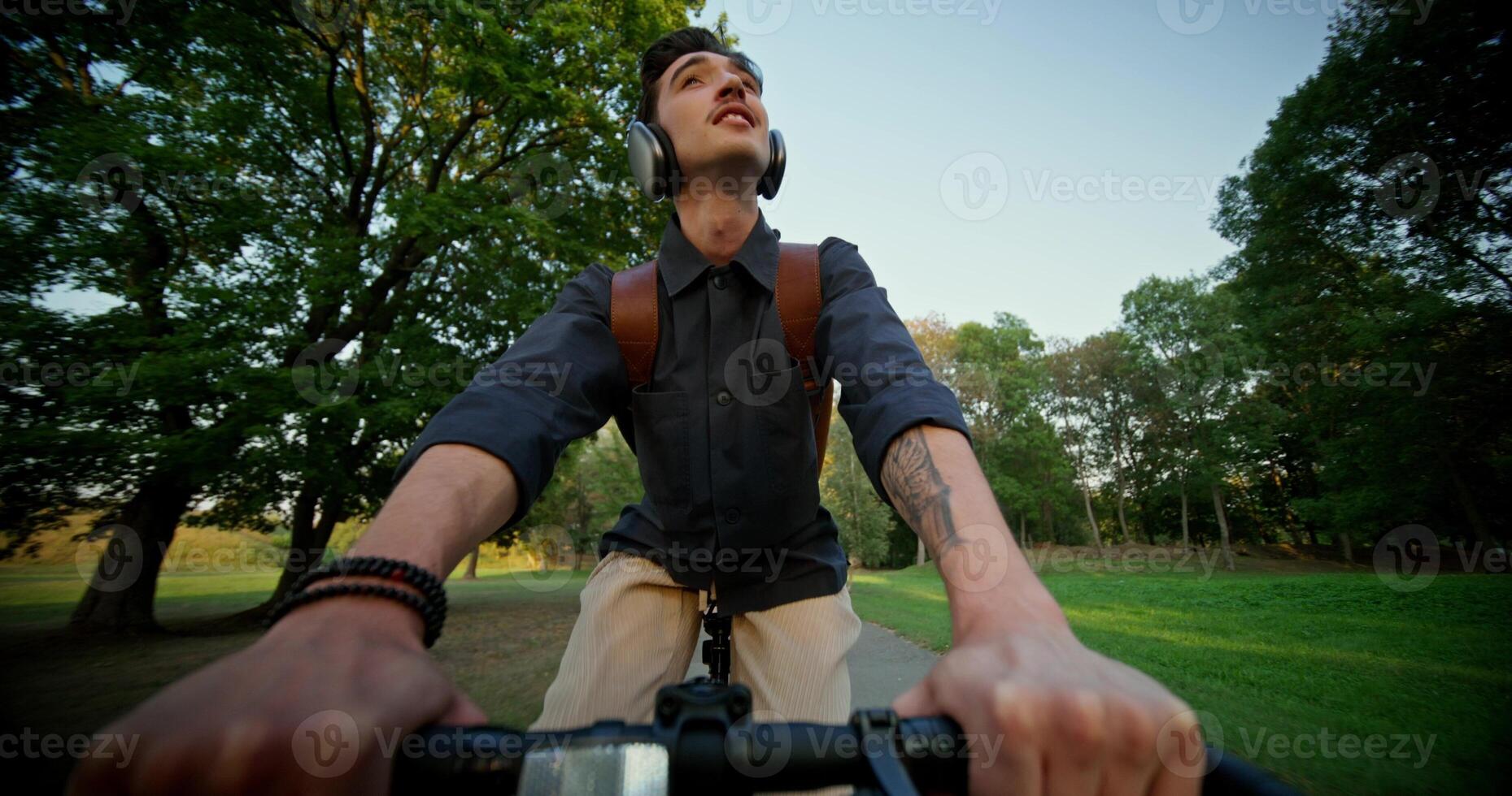 A young man with headphones and a backpack on his shoulders rides a bicycle in a park. Front view. Summer day photo