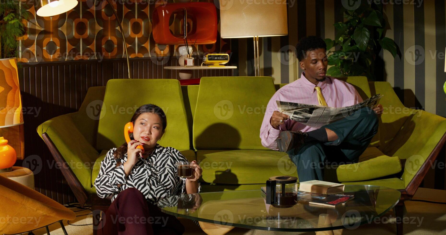A man is reading a newspaper with a concentrated look while sitting on the couch at home. His wife is on the phone sitting on the floor next to him. Retro interior photo