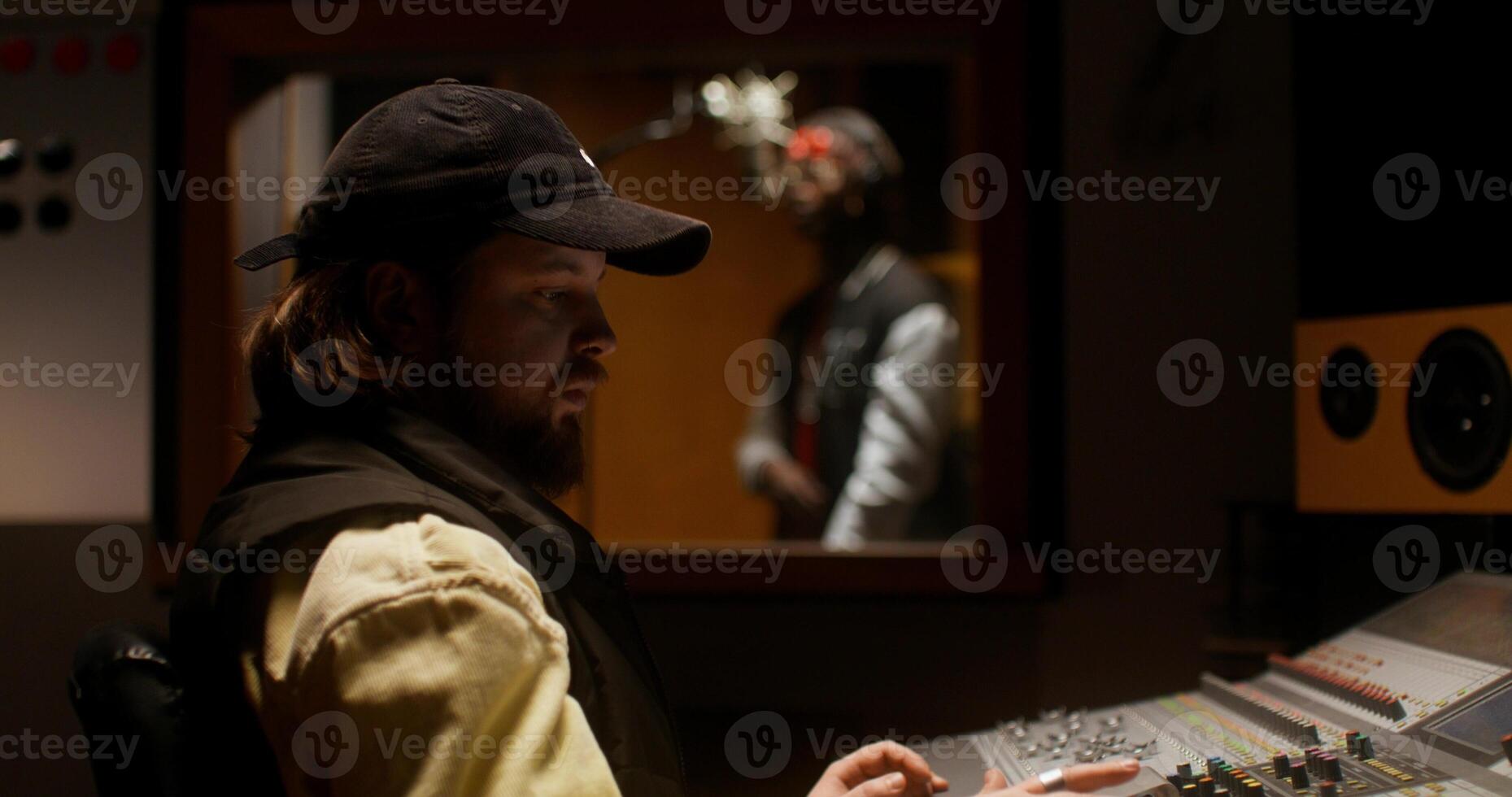A sound engineer uses an audio mixer while recording a song in the studio. The rapper sings in the room behind the glass photo