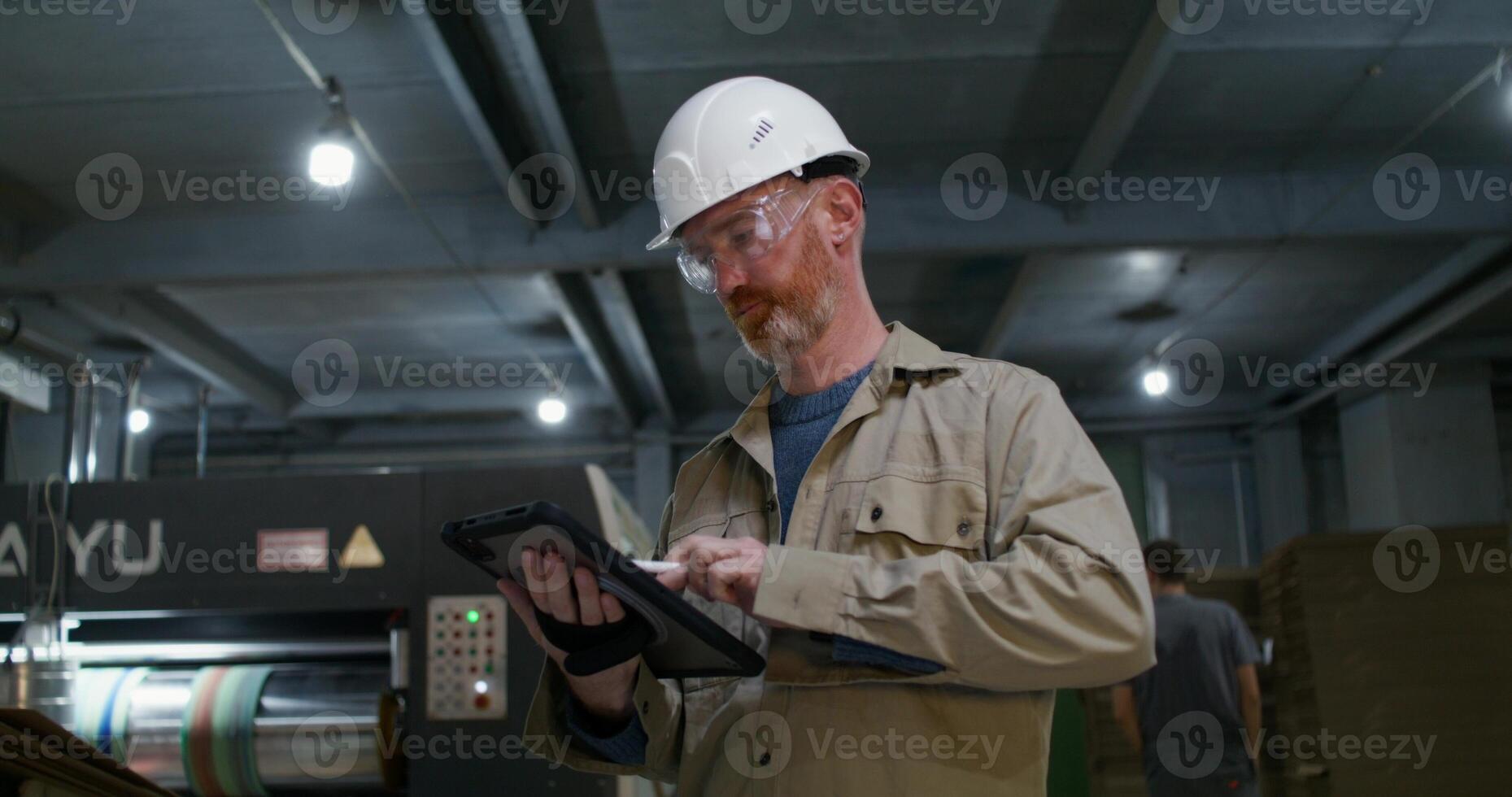 A man in a protective helmet and glasses uses a tablet while working at a factory. He stands near production machine photo