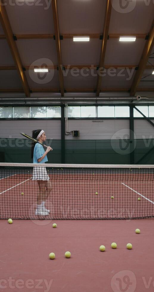 A young woman of Asian appearance in a short skirt comes to the tennis court with a tennis racket in her hands and leaning against a stretched tennis net posing looking at the camera, vertical photo
