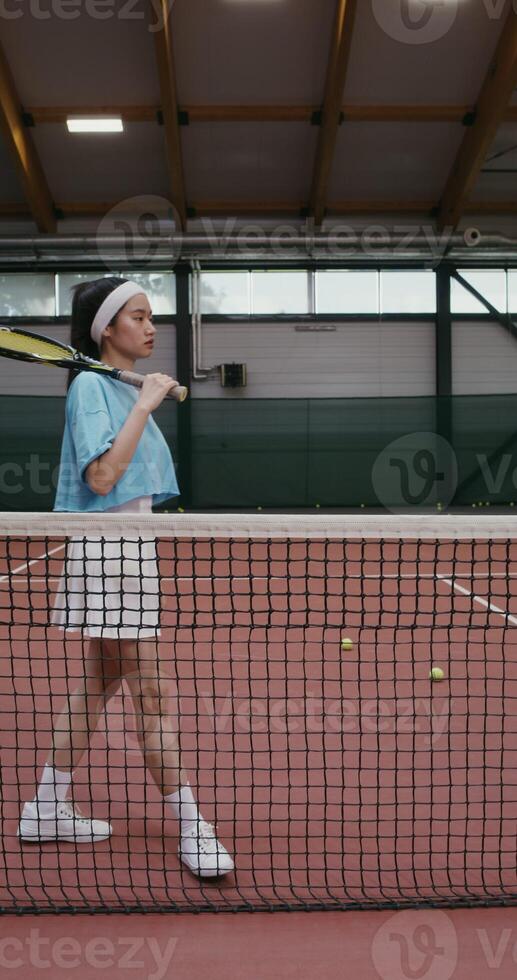 A young woman of Asian appearance in a short skirt comes to the tennis court with a tennis racket in her hands and leaning against a stretched tennis net posing looking at the camera photo