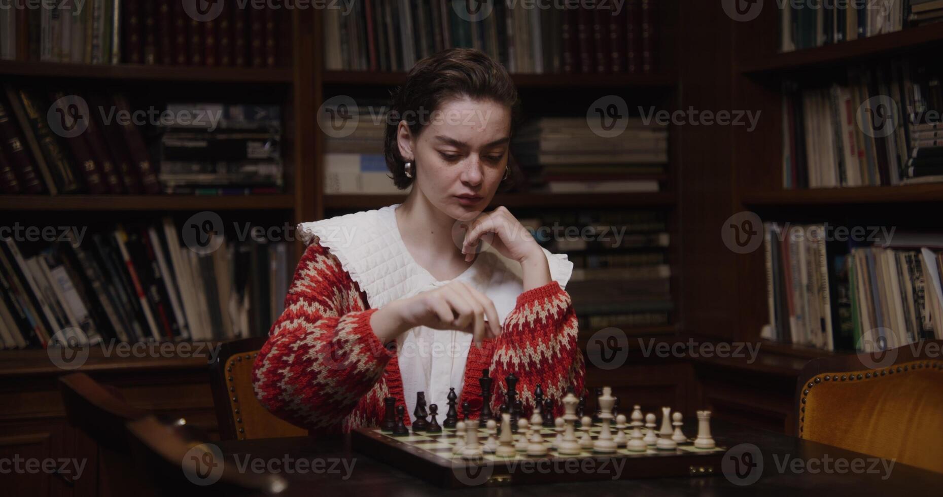 A woman of European appearance plays a chess game alone, thinking long concentratedly between moves, sitting at a table in the library photo