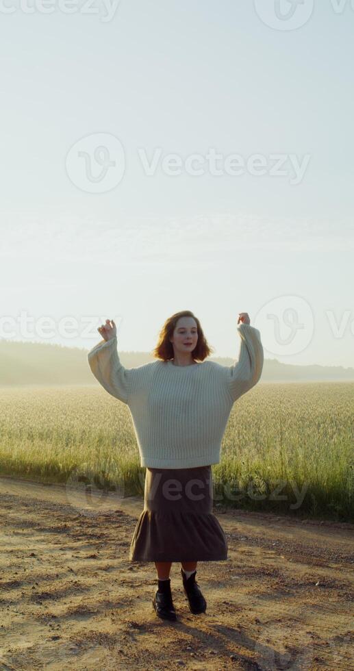 A beautiful young woman, dressed in a warm sweater, laughs and spins around, dancing on gravel road near an agricultural field, enjoying the warm rays of the rising sun photo