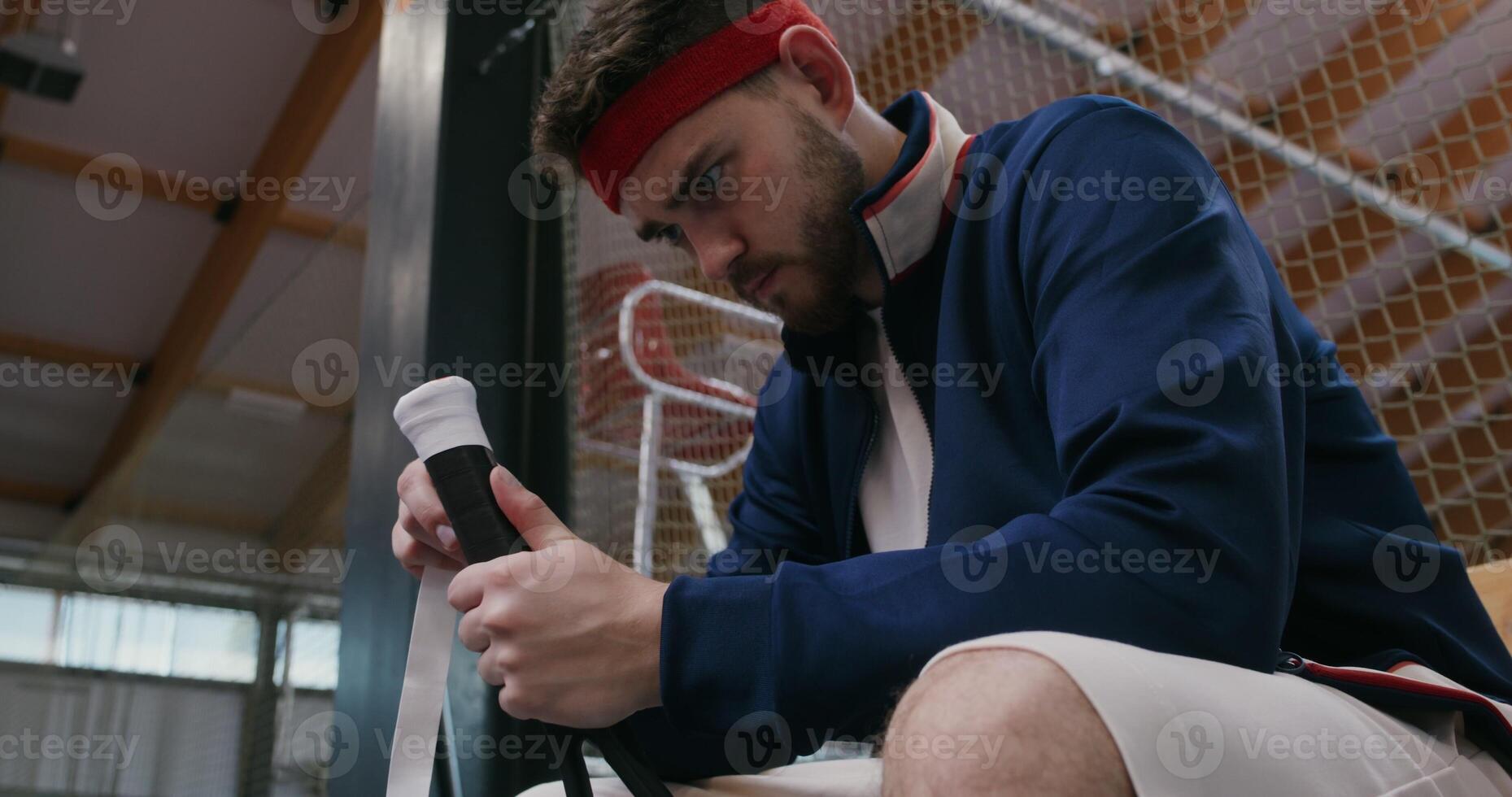 A young man of European appearance wraps the handle of a tennis racket with a special tape for better grip on the hand, sitting on a bench near the tennis court photo