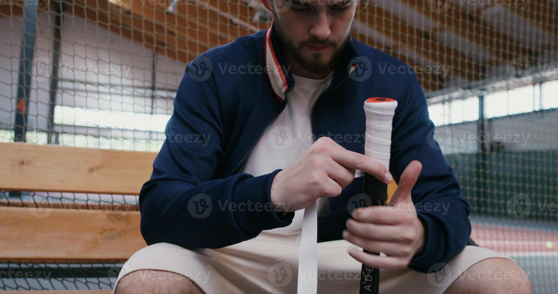 A young man of European appearance wraps the handle of a tennis racket with a special tape for better grip on the hand, sitting on a bench near the tennis court, preparing for a tennis match, close-up photo