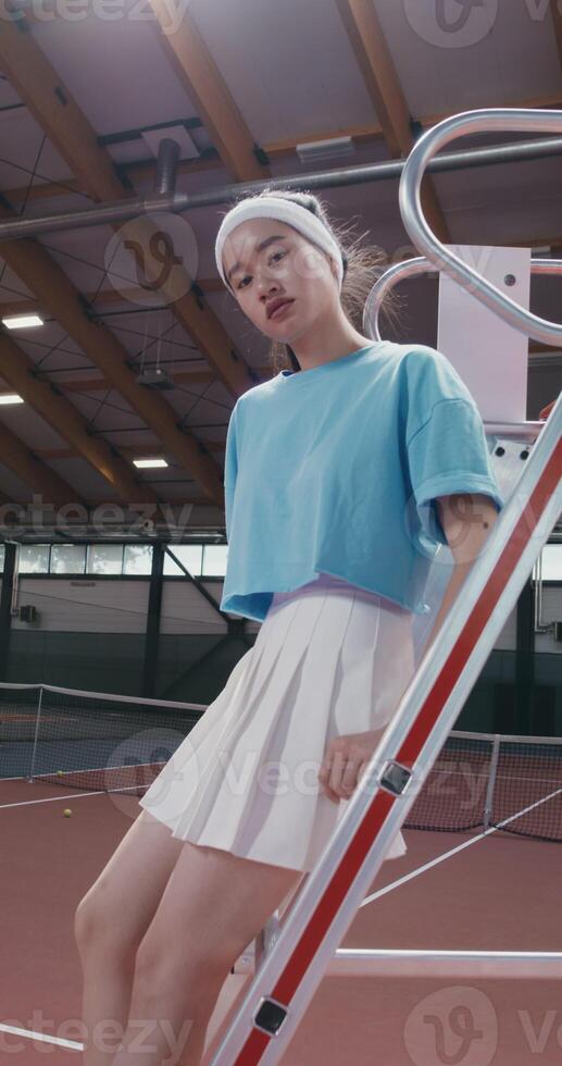 A woman tennis player of Asian appearance in a short skirt sits on the stairs of the referee's chair in the middle of an indoor tennis court and looks at the camera with a serious look photo