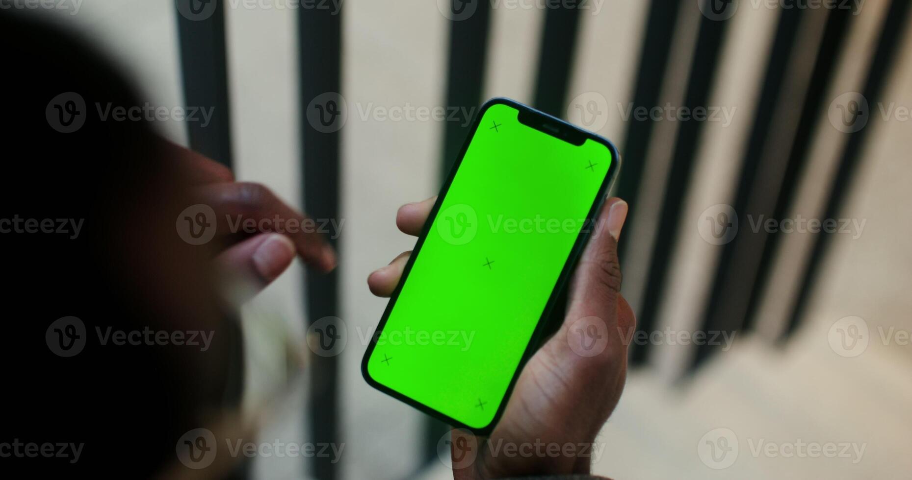 A mobile with a green screen in the hand of a man of African American appearance scrolling the screen, close-up, filming from behind. Copy space, mockup photo