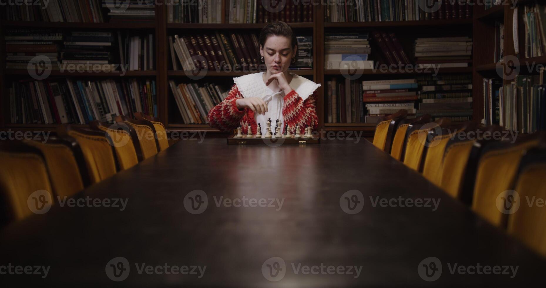 A young woman of European appearance plays a chess game alone, thinking long concentratedly between moves, sitting at a long table photo
