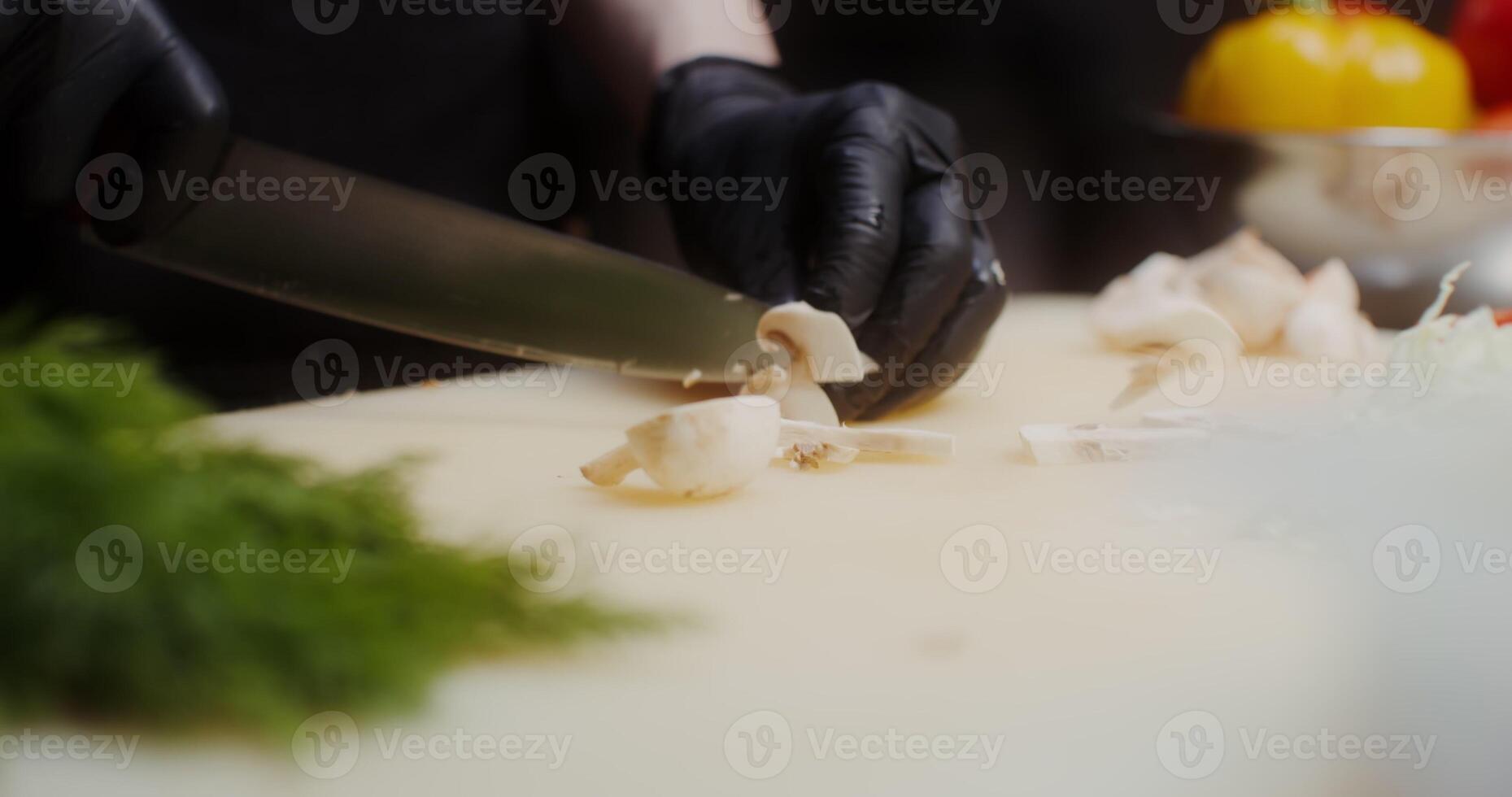A woman in a chef's uniform with disposable black gloves on her hands cuts tomato standing in the restaurant kitchen. Close photo