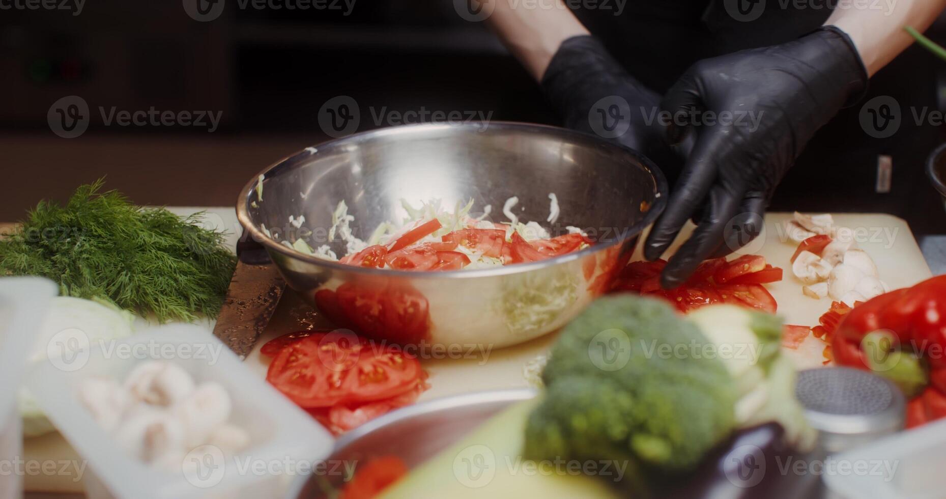 A woman in a chef's uniform with disposable black gloves on her hands puts chopped vegetables in a bowl standing in the restaurant kitchen, close photo
