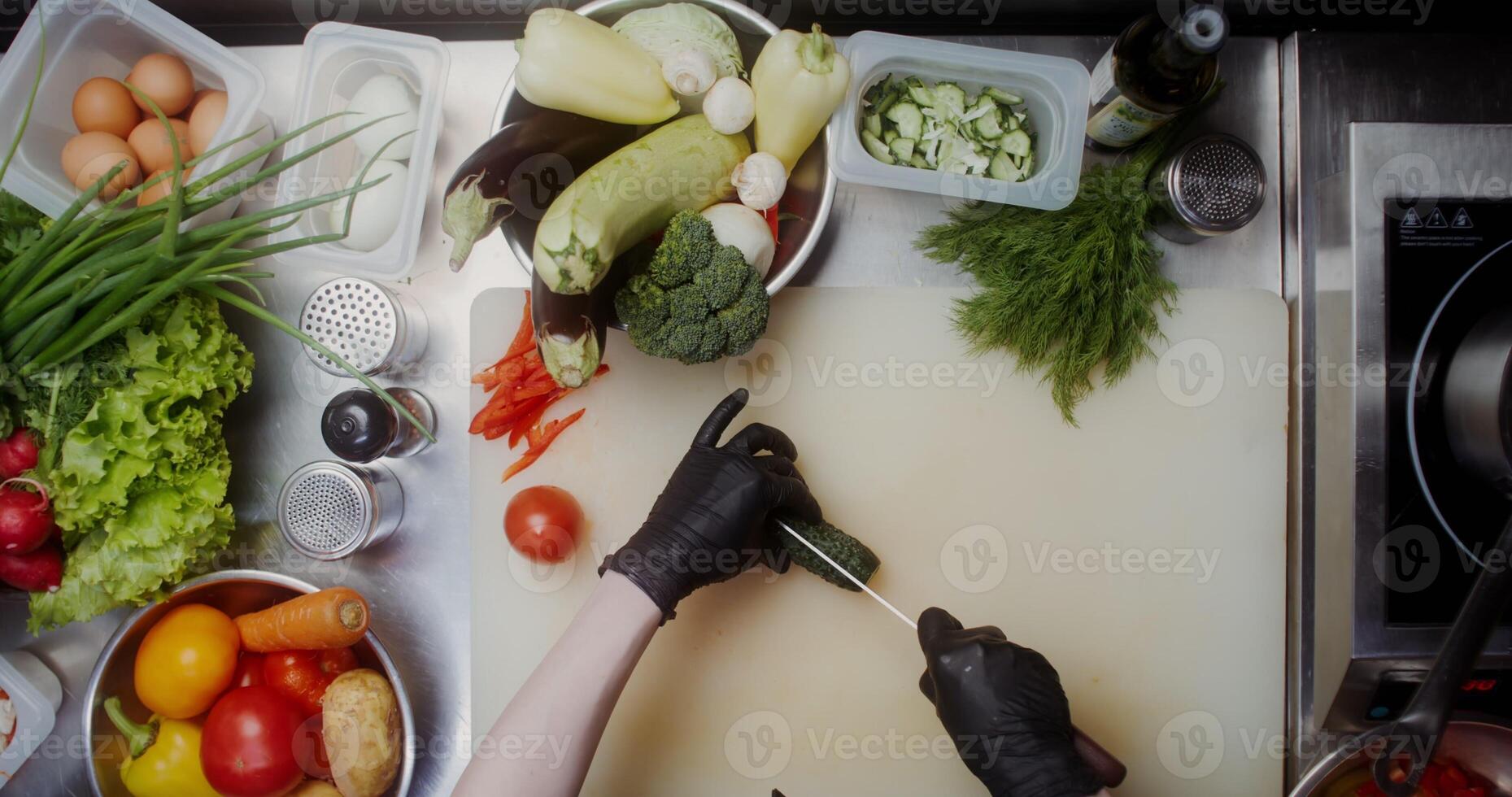 A woman in disposable black gloves cuts cucumbers standing near a table full of vegetables, close photo