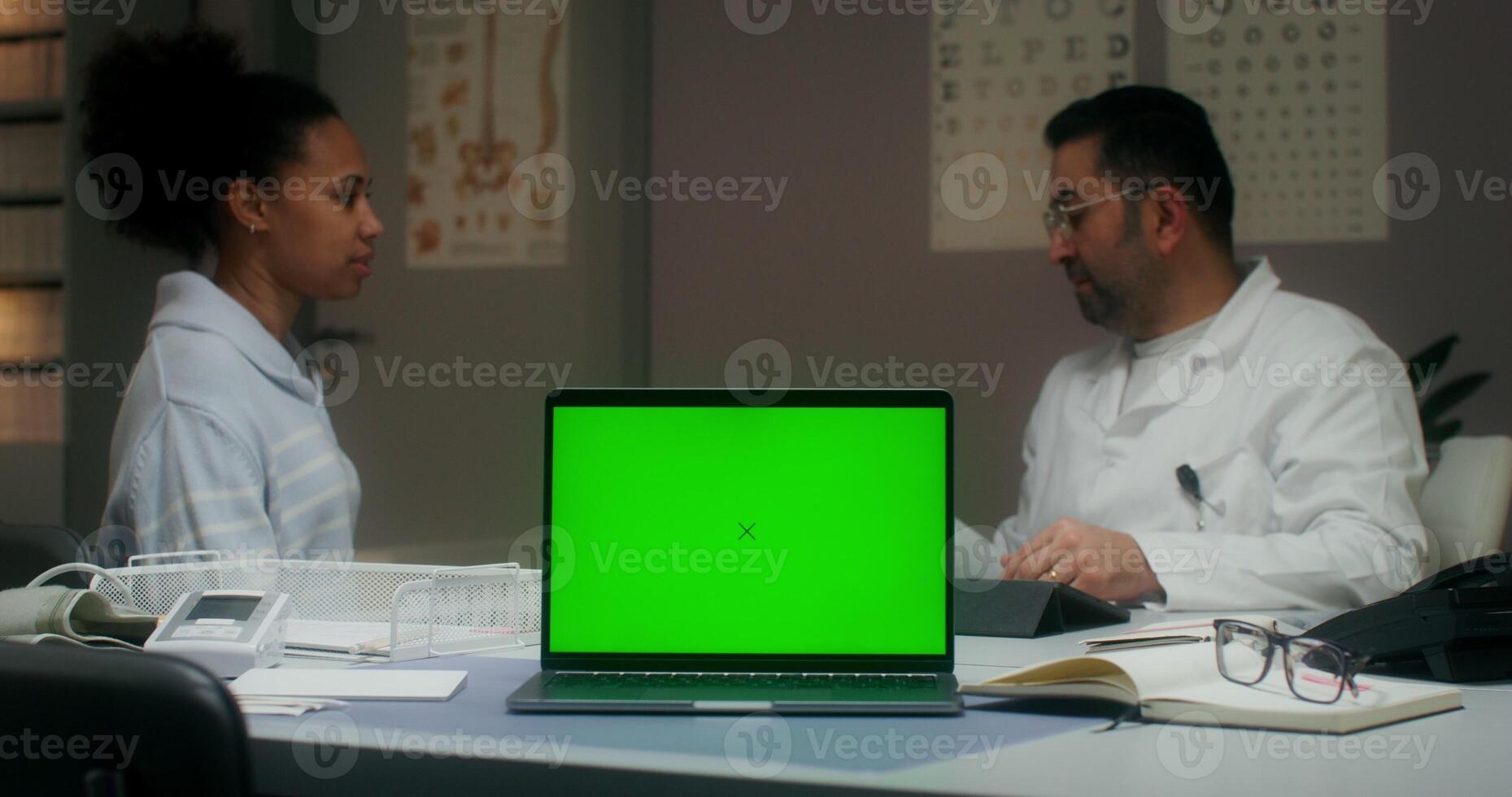 A laptop with a green screen is on the table in the medical office. The doctor measures the temperature of a patient photo