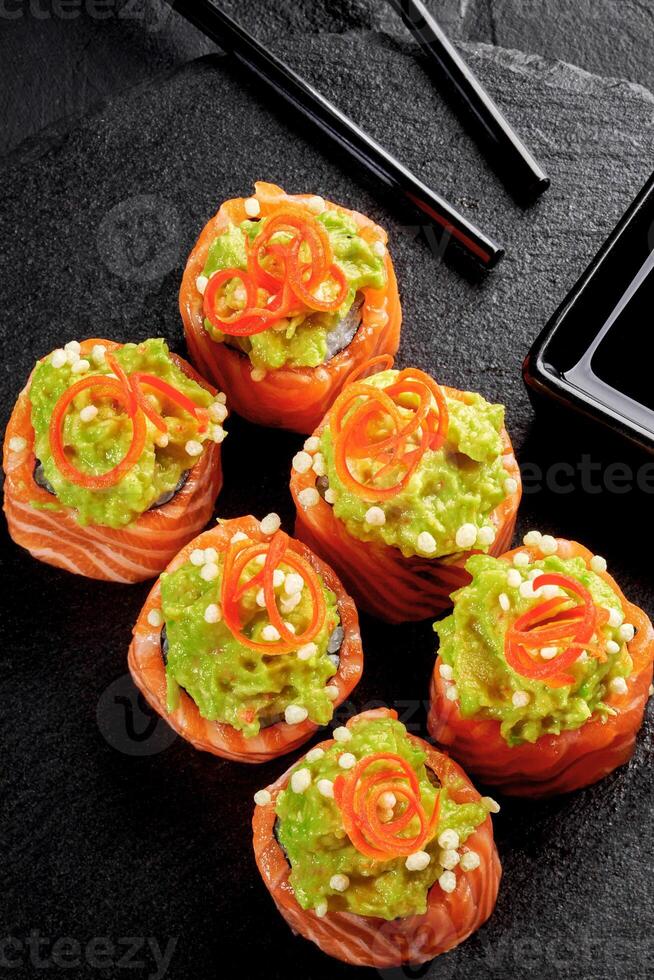 Delicious uramaki sushi rolls covered with salmon and guacamole, decorated with chili pepper and sesame seeds, resting on black stone plate photo