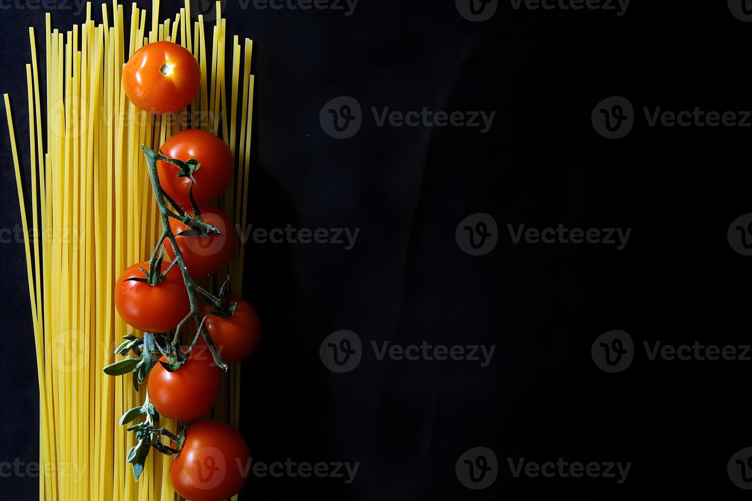 A bunch of tomatoes on the vine rests next to spaghetti against black background photo