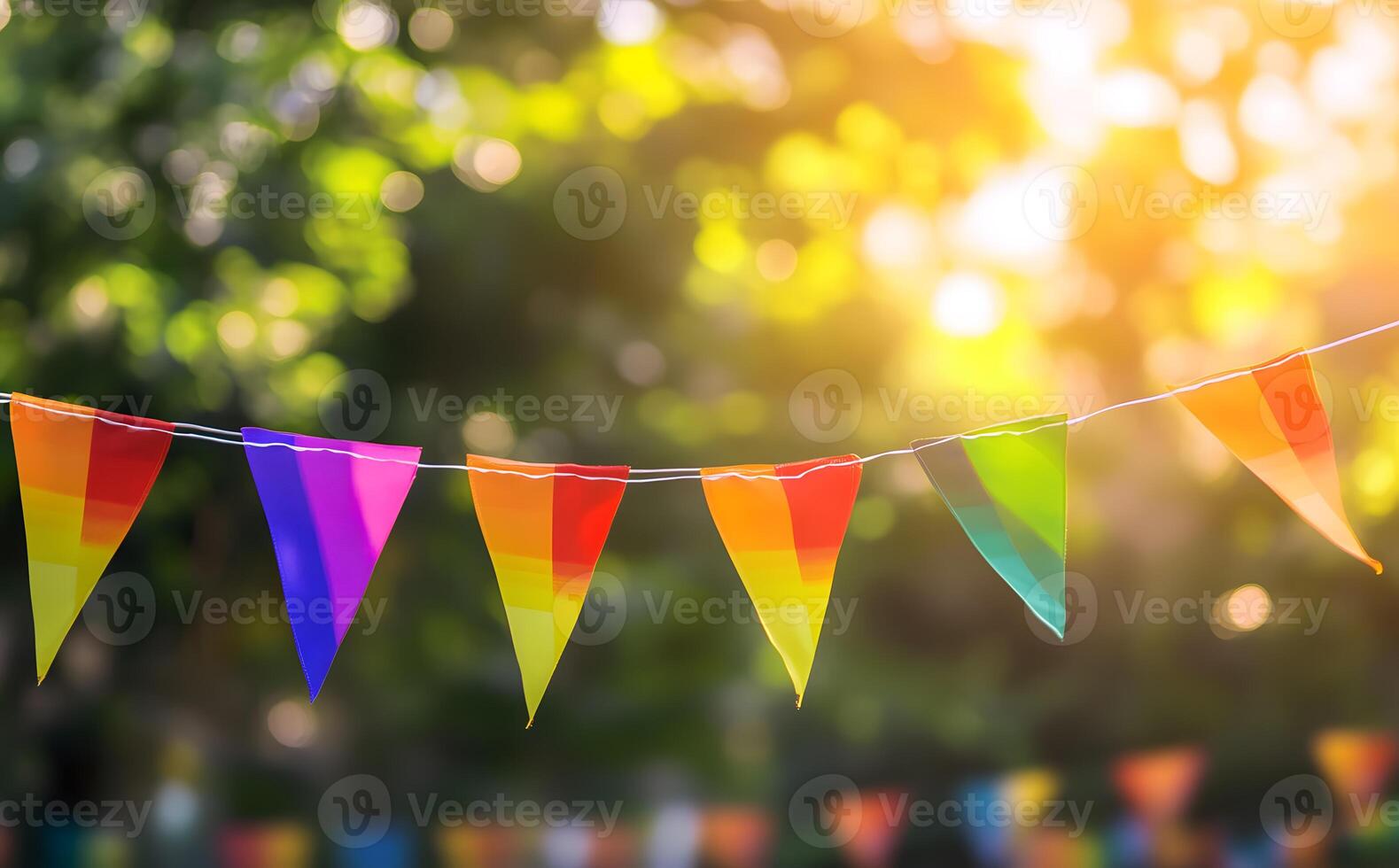 String of rainbowcolored pennant flags hangs against a blurred, sunlit, and green background photo