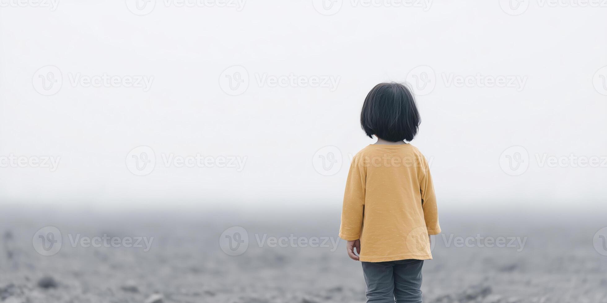 Young girl with short black hair wearing a yellow shirt stands alone in a vast, foggy landscape, evoking feelings of solitude and contemplation in a serene environment photo
